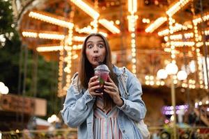 foto al aire libre de una encantadora mujer joven con el pelo largo y castaño mirando a la cámara con los ojos bien abiertos y contrayendo la frente, bebiendo limonada mientras camina por el parque de atracciones