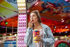 Portrait of attractive young brunette woman with sunglasses on her head eating popcorn while walking in amusement park, looking aside with interest photo