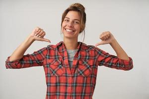 joven mujer feliz y hermosa con peinado de moño con camisa a cuadros y camiseta, posando sobre fondo blanco con las manos levantadas, apuntándose con los pulgares a sí misma y sonriendo ampliamente foto