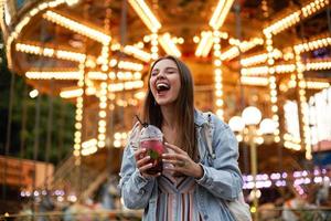 Outdoor portrait of joyful young pretty brunette female in casual clothes posing over amusement park with closed eyes and broad smile, holding cup of lemonade in hands photo