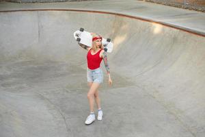 foto al aire libre de una joven rubia tatuada parada sobre un parque de patinaje con jeans cortos y top rojo, sosteniendo una tabla sobre sus hombros y mirando a un lado con cara tranquila