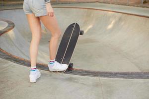 Cropped outdoor photo of slim female in jeans shorts posing over skateboard on summer warm weather, going to ride board in stunt park