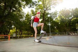 Young pretty blonde long haired woman with tattoos standing over green park on sunny warm day, standing on the edge of concrete wall, going to learn skating photo