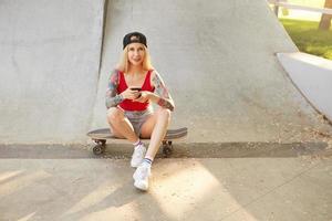 Young beautiful blonde tattooed female in baseball cap looking to camera in positiv mood, sitting on skateboard over city park with smartphone in hands photo