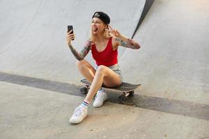 Funny outdoor shot of lovely young tattoed long haired female posing over concrete wall while sitting on board, making selfie with her smartphone, raising hand with victory sign and showing tongue photo