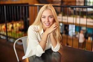 Indoor portrait of beautiful young long haired blonde woman posing over cafe interior, looking at camera with charming smile and leaning head on crossed arms photo