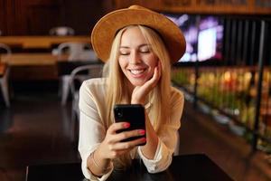 Indoor close-up of charming blonde young female in white shirt and brown hat sitting over cafe interior, holding mobile phone in hand and looking at screen with cheerful smile photo