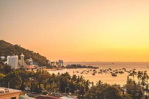 vista aérea de la ciudad de vung tau con hermosa puesta de sol y tantos barcos. vista panorámica costera de vung tau desde arriba, con olas, costa, calles, cocoteros y montaña tao phung en vietnam. foto