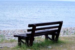 Brown wooden bench in the park. Summer sunny day. Green grass and trees. Resting and relaxing area. Empty bench for sitting. Wood exterior material. Beautiful bench near sea or lake photo