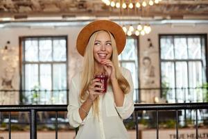 Indoor shot of happy young blonde woman with blue eyes standing over restaurant interior, drinking lemonade with straw and looking aside with dreamy smile photo