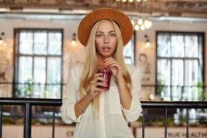 Indoor shot of beautiful blonde long haired woman with blue eyes posing over restaurant interior, drinking lemonade with straw, wearing hat and shirt