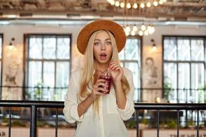 Surprised young blonde woman in wide hat posing over cafe interior, looking to camera with wide mouth opened and holding cup of lemonade in her hands photo