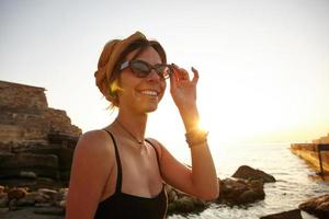 Outdoor photo of beautiful young dark haired female with casual hairstyle wearing black swimwear and vintage sunglasses, posing over seaside on early summer morning