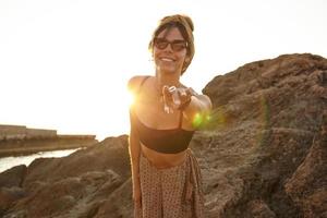 Attractive young dark haired woman in mustard color headband and sunglasses posing over sunrise view on early morning, raising hand and asking happily to follow her photo