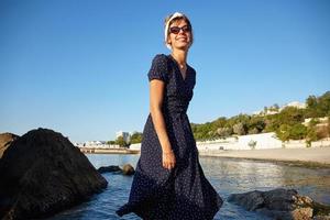 foto al aire libre de una atractiva joven de cabello oscuro vestida de verano posando sobre la playa en un día cálido y soleado, de buen humor y sonriendo alegremente
