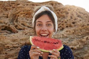 Outdoor photo of charming young dark haired woman in headband standing over big stone and holding piece of watermelon in her hands, contracting forehead with wide mouth opened