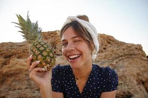 primer plano de una joven y atractiva mujer de cabello oscuro con diadema mirando a la cámara alegremente y guiñando un ojo, sosteniendo piña fresca en la mano, usando ropa informal foto