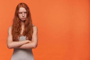 Indoor photo of young red haired lovely woman looking aside and frowning her face in displeasure, keeping arms folded while standing over orange background in casual clothes