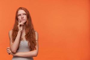 Cute young pensive female with wavy foxy hair lond hair looking upwards while thinking, isolated over orange background, wearing casual grey shirt, holding hand on her face photo