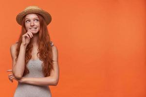 Lovely young wavy lond haired foxy woman in grey shirt and straw hat holding her chin, looking aside with dreamy face and biting underlip, isolated over orange background photo