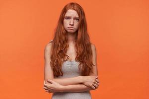 mujer joven y bonita ofendida con el pelo ondulado y astuto posando sobre un fondo naranja, mirando la cámara con cara triste, usando ropa informal, manteniendo las manos dobladas foto