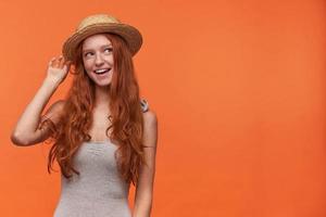 Positive young beautiful woman with wavy foxy hair posing over orange background, wearing grey shirt and boater hat, looking cheerfully aside and raising hand to straighten headdress photo