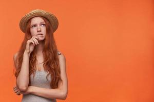 Puzzled young pretty woman with wavy foxy hair leaning her face on raised hand and looking aside doubtfully, posing over orange background in casual clothes photo