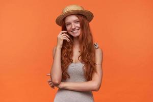 foto de estudio de una joven y bonita mujer de pelo rojo con ropa informal y sombrero de navegante, parada sobre un fondo naranja, mirando a la cámara con una sonrisa encantadora y tocando su cabello ondulado