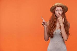 Dazed attractive young lady with wavy foxy hair posing over orange background in casual clothes, raising hand and pointing aside with index finger, keeping palm on her mouth and raising eyebrows photo