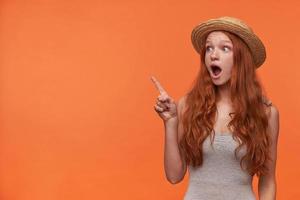 Surprised attractive young female with wavy foxy hair standing over orange background with raised index finger, looking aside pop-eyed with wide opened mouth photo