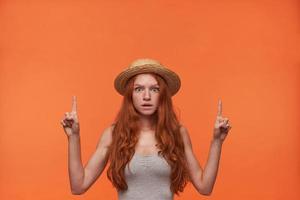 Indoor shot of young pretty lady with wavy foxy hair posing over orange background with wide eyes opened, pointing up with forefingers and looking to camera seriously, wearing casual clothes photo