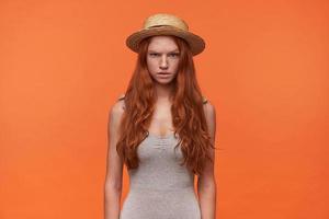 atractiva joven mujer de pelo largo y astuta posando sobre un fondo naranja con ropa informal con las manos hacia abajo, mirando a la cámara con cara seria y levantando una ceja foto