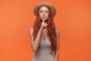 retrato de una joven encantadora con el pelo ondulado y astuto levantando el dedo índice a los labios, pidiendo guardar silencio, posando sobre un fondo naranja con camisa gris y sombrero de paja foto