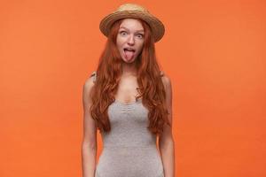 Funny indoor shot of joyful pretty young redhead woman with long hair, looking to camera and showing tongue, posing over orange background in casual clothes photo