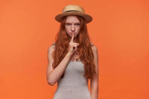 foto de estudio de una hermosa joven mujer de pelo largo y astuto levantando el dedo índice en un gesto silencioso, mirando a la cámara con cara seria y pidiendo silencio, aislada sobre fondo naranja