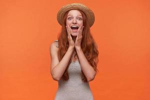 Horizontal shot of attractive young redhead woman with lond hair standing over orange background in grey shirt and straw hat, looking to camera with wide cheerful smile and holding hands on her cheeks photo