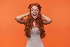 Studio shot of lovely overjoyed young female with wavy foxy lond hair posing over orange background, looking to camera cheerfully and holding her head with, being happy to madness photo