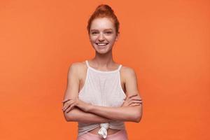 Indoor photo of beautiful young female with foxy hair standing over orange background, keeping hands folded and looking at camera with charming smile, wearing casual clothes