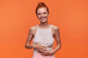 Cheerful lovely young lady with foxy hair combed in bun wearing white top, standing over orange background with hands on her body, smiling to camera widely photo