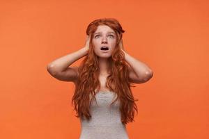 joven mujer abrumada de cabello bastante largo sosteniendo su cabeza con las manos y mirando hacia arriba con la cara conmocionada, posando sobre un fondo naranja con ropa informal foto