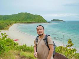 mochilero gordo asiático se encuentra en el punto de vista en la parte superior de la isla con idílica playa, océano y cielo azul en tiempo de vacaciones, samae san island chonburi tailandia, concepto de verano foto