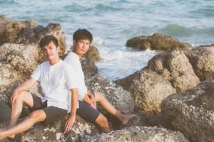 retrato homosexual joven pareja asiática sentada abrazada en roca o piedra en la playa en verano, turismo gay de asia para el ocio y relajarse con felicidad en vacaciones en el mar, concepto legal lgbt. foto