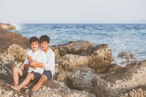 Homosexual portrait young asian couple sitting hug together on rock or stone in the beach in summer, asia gay going tourism for leisure and relax with happy in vacation at sea, LGBT legal concept. photo