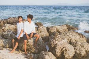 retrato homosexual joven pareja asiática sentada abrazada en roca o piedra en la playa en verano, turismo gay de asia para el ocio y relajarse con felicidad en vacaciones en el mar, concepto legal lgbt. foto