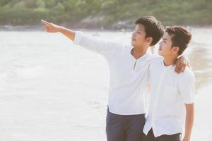 Homosexual portrait young asian couple standing pointing something together on beach in summer, asia gay going sea for leisure with romantic and happy in vacation at sea, LGBT with legal concept. photo