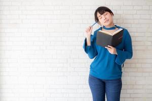 Beautiful asian woman smiling standing thinking and writing notebook on concrete cement white background at home, girl homework on book, education and lifestyle concept. photo