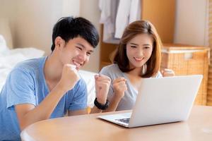 Beautiful portrait young asian couple working laptop with smile and happy sitting in bedroom, man and woman using notebook computer with excited and satisfied, business and success concept. photo