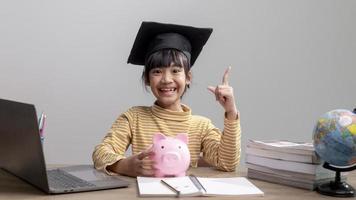 Asian little girl wearing a graduation cap with a pink piggy bank, Saving money, investment the future, photo