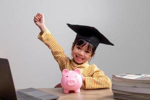 niña asiática con una gorra de graduación con una alcancía rosa, ahorrando dinero, invirtiendo en el futuro, foto