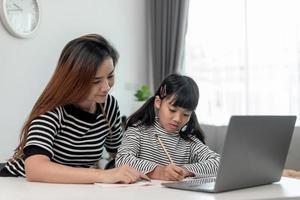 Asian little young girl kid learning online class at home with mother. Preschool child use laptop computer do homework, homeschool from school teacher by digital remote internet with support from mom. photo